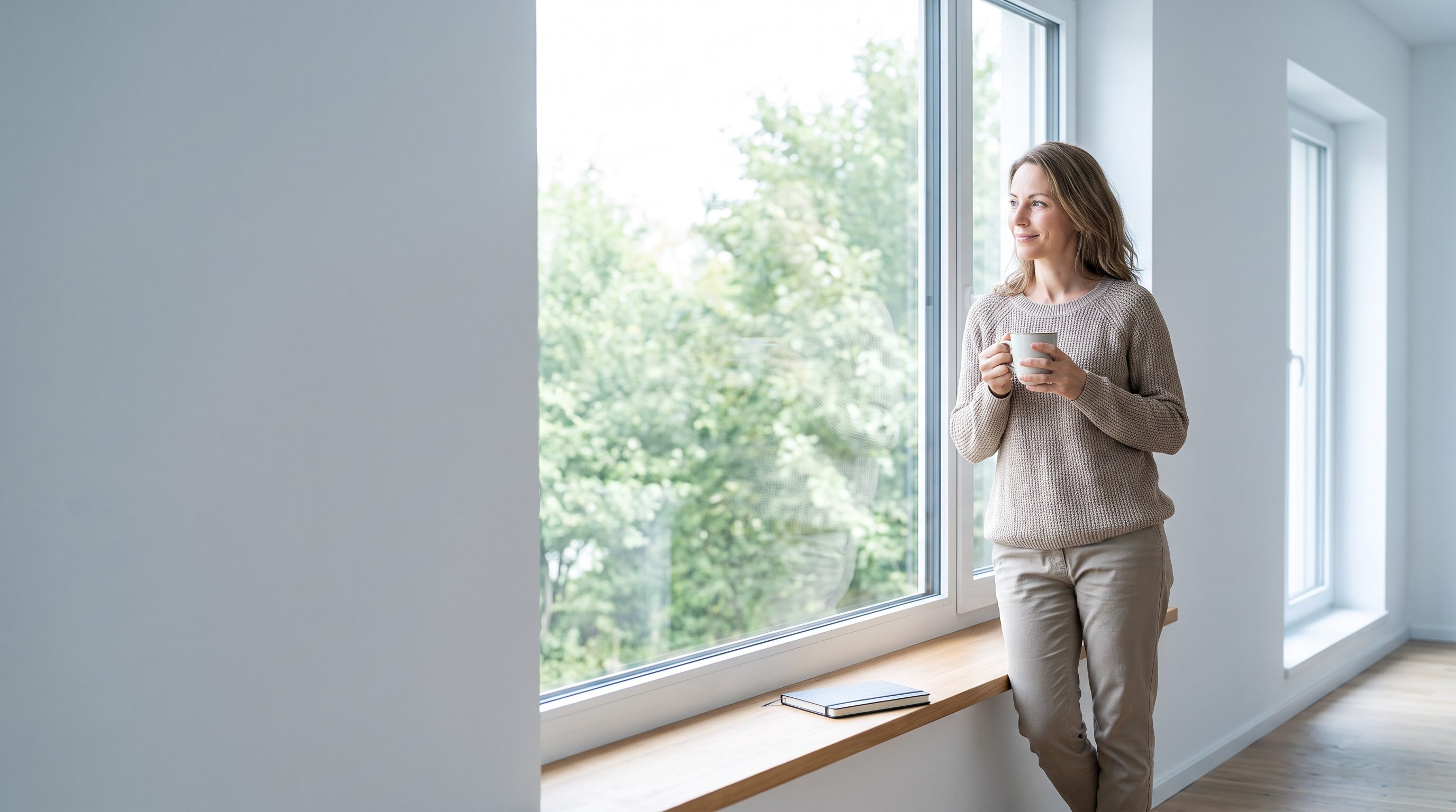 Frau steht mit einer Tasse Kaffee am Fenster und schaut ins Grüne