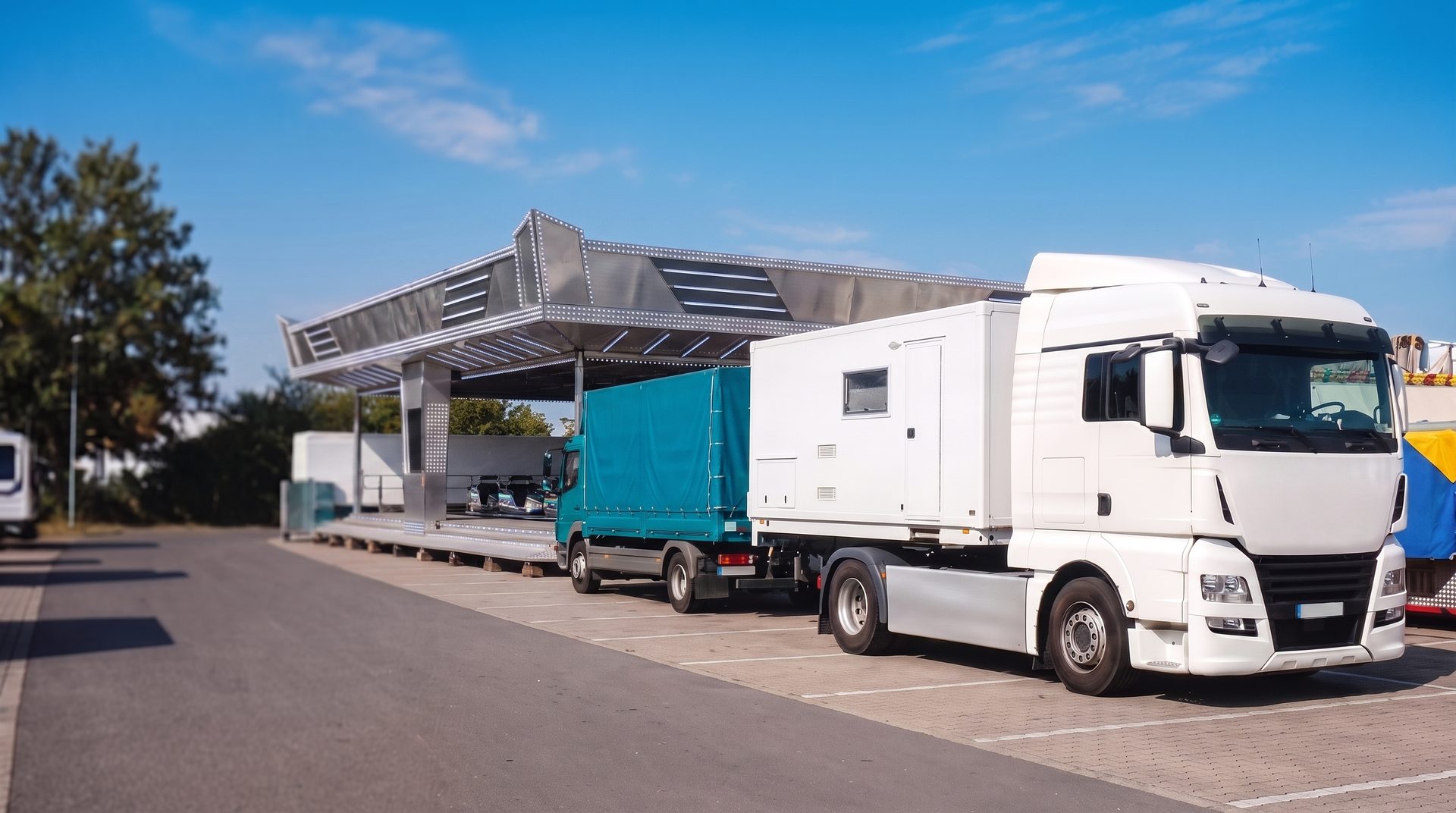 Moderner Schausteller-Fuhrpark an einem hellen Sommertag, weißer Sattelzug mit Wohnwagen und Pritschen-LKW mit türkiser Plane vor einem modernen Fahrgeschäft auf dem Festplatz
