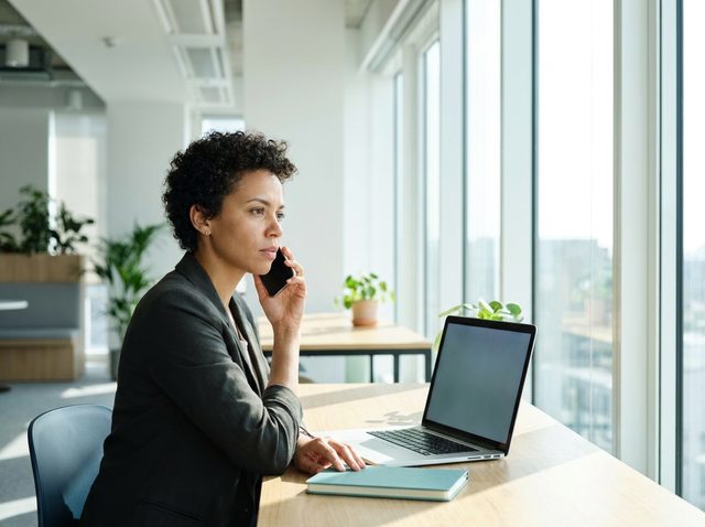 Frau am Schreibtisch im Büro telefoniert, der Bildschirm ihres Laptops ist dunkel