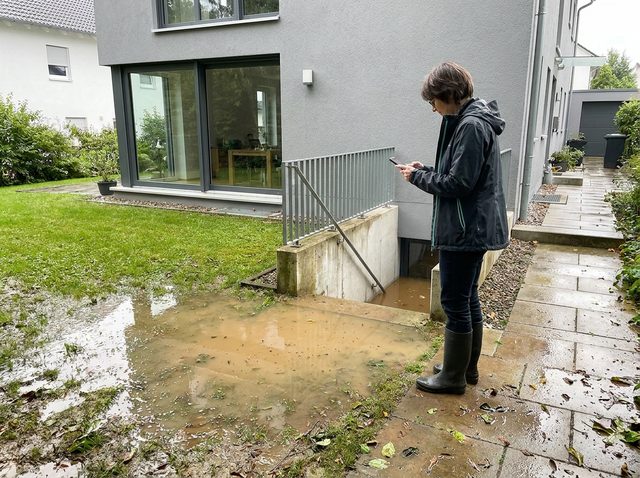 Überschwemmter Rasen und Lichtschacht an einem Wohnhaus, eine Frau in Gummistiefeln macht Aufnahmen